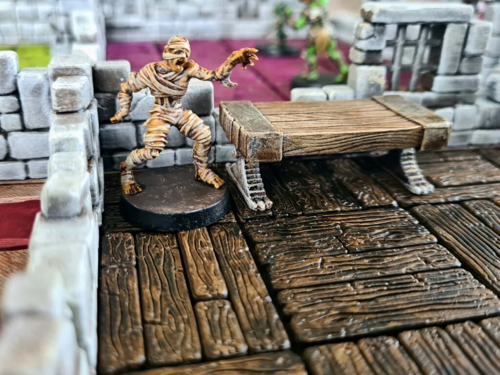 Figurine de momie HeroQuest sur un sol en parquet de bois 3D détaillé avec un établi d'alchimiste en bois.
